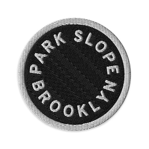 May include: Embroidered patch with the text "BROOKLYN PARK SLOPE" in white, encircling a black center. The patch has a silver-gray border and is circular in shape.