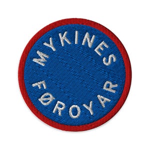 May include: Circular embroidered patch with a blue centre and red border. The text "MYKINES FØROYAR" is embroidered in white around the blue centre. The patch is likely for clothing or accessories.