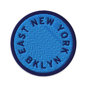 May include: A circular embroidered patch featuring a bright blue centre with the dark blue text EAST NEW YORK BKLYN arranged in a circle. The patch has a thick, dark blue stitched border that frames the light blue interior design.