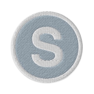 Puede incluir: Parche bordado con la letra "S" blanca sobre un óvalo azul claro. El óvalo está rodeado por un borde blanco, creando un efecto de capas. El parche tiene una apariencia texturizada, sugiriendo un material de tela.