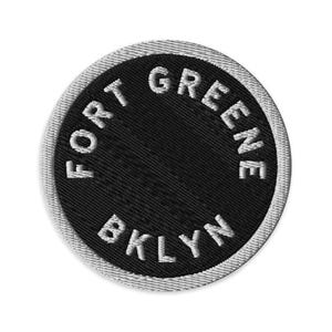 May include: A circular embroidered patch featuring a black center with the white text FORT GREENE BKLYN stitched in a circular pattern along the edge. The patch is finished with a thick, light gray stitched border around the entire circumference.