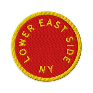 Pode incluir: Um emblema bordado circular com um fundo vermelho vibrante e uma borda amarela espessa. O texto LOWER EAST SIDE NY é costurado com fio amarelo em um padrão circular ao longo da borda interna do emblema, centralizado no campo vermelho.