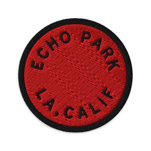Può includere: Una toppa ricamata circolare con uno sfondo rosso vibrante e un bordo nero spesso. Il testo "ECHO PARK" è cucito in nero lungo la curva superiore, e "L.A., CALIF." è cucito in nero lungo la curva inferiore della toppa.