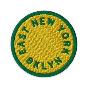 Puede incluir: Un parche bordado circular con un centro amarillo brillante y un borde verde grueso. El texto "EAST NEW YORK BKLYN" está bordado con hilo verde, curvándose a lo largo del borde interior del círculo amarillo en una fuente negrita sin remates.