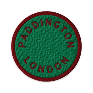 May include: A circular embroidered patch featuring a vibrant green centre surrounded by a dark red border. The text PADDINGTON LONDON is stitched in dark red capital letters, following the circular curve of the patch's outer edge.