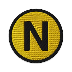 May include: Embroidered patch featuring a large black letter "N" centered on a yellow circle. The yellow circle has a textured appearance and is surrounded by a black border. The patch is circular and likely designed to be sewn or ironed onto fabric.