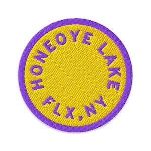 Könnte beinhalten: Ein kreisförmiger bestickter Aufnäher mit einer leuchtend gelben Mitte und einem violetten Rand. Der Text HONEOYE LAKE FLX, NY ist in violettem Garn gestickt und folgt dem kreisförmigen Rand des Aufnähers mit einer sauberen Textur.
