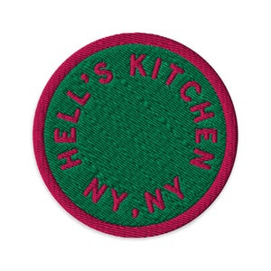 Puede incluir: Un parche bordado circular con un centro verde oscuro y un borde rosa vibrante. El texto HELL'S KITCHEN NY, NY está bordado con hilo rosa en un patrón circular alrededor del borde interior del parche, creando un contraste llamativo.