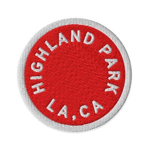 May include: A circular embroidered patch featuring a vibrant red center with the white text HIGHLAND PARK LA, CA stitched in a circular pattern. The patch is finished with a thick, white stitched border around the entire outer edge.