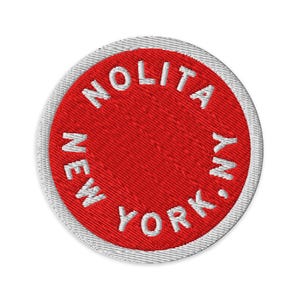 May include: A circular embroidered patch featuring a vibrant red background with a white border. The text NOLITA NEW YORK, NY is stitched in white capital letters following the circular edge of the patch, creating a clean and bold design.