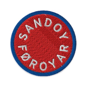 Puede incluir: Parche bordado con centro rojo y borde azul. El parche presenta texto blanco en un patrón circular que dice "SANDØYAR FØROYAR". El diseño es circular y tiene una apariencia texturizada.