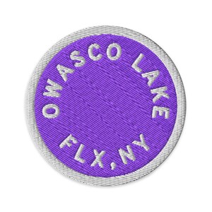 Könnte beinhalten: Ein kreisförmiger gestickter Aufnäher mit einer leuchtend violetten Mitte und weißem gesticktem Text, der OWASCO LAKE FLX, NY in einem kreisförmigen Muster anzeigt. Der Aufnäher ist mit einem weißen gestickten Rand versehen.