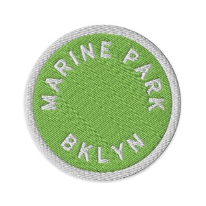 May include: A circular embroidered patch featuring a bright lime green centre with a white border. The text MARINE PARK is stitched in white along the top curve, and the text BKLYN is stitched in white along the bottom curve of the patch.