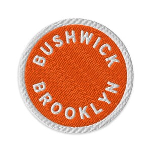 May include: Embroidered patch with the words "Bushwick Brooklyn" in white, arched around a bright orange circle. The patch has a white border and is circular in shape. The design is simple and bold.