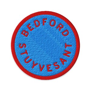 Puede incluir: Un parche redondo bordado con un centro azul y un borde rojo. El texto "BEDFORD STUYVESANT" está bordado en rojo alrededor del centro azul. El parche tiene forma circular.