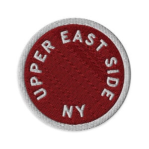 Könnte beinhalten: Ein kreisförmiger bestickter Aufnäher mit einer tiefroten Mitte und dem weißen Text UPPER EAST SIDE NY, der kreisförmig entlang des Randes gestickt ist. Der Aufnäher hat einen dicken, hellgrau gestickten Rand, der das Design einfasst.