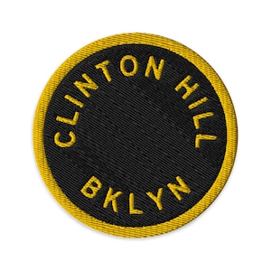 Puede incluir: Un parche bordado circular con fondo negro y un borde amarillo grueso. El texto CLINTON HILL está arqueado en la mitad superior y la abreviatura BKLYN está centrada a lo largo del borde inferior, todo bordado con hilo de color amarillo.