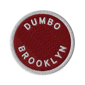 May include: A circular embroidered patch with a deep red center and a white border. The text DUMBO is stitched in white along the top curve, and the text BROOKLYN is stitched in white along the bottom curve of the red circular field.