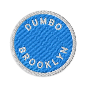 May include: A circular embroidered patch featuring a bright blue center with the white text DUMBO BROOKLYN arched along the top and bottom. The patch has a thick, light gray stitched border that frames the blue circular design completely.