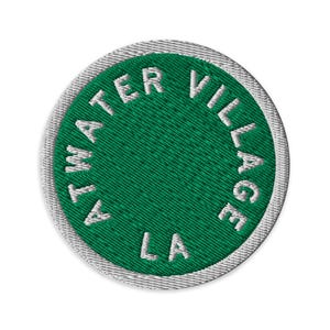 May include: A circular embroidered patch featuring a deep green background with a white border. The text ATWATER VILLAGE LA is stitched in white capital letters following the circular edge of the patch, creating a clean and distinct design.