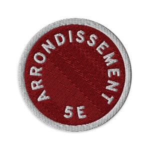 May include: A circular embroidered patch featuring a deep red center and a white border. The white text "ARRONDISSEMENT 5E" is stitched in a circular pattern along the inner edge of the red field, creating a clean and distinct graphic design.