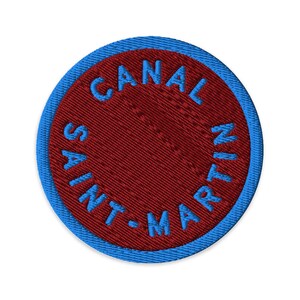 May include: A circular embroidered patch featuring a deep red center and a bright blue border. The text CANAL SAINT-MARTIN is stitched in bright blue thread, following the circular curve of the patch along the inner edge of the blue border.