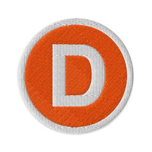 May include: Embroidered patch featuring a white letter "D" inside an orange circle. The circle is surrounded by a white border. The patch has a textured, stitched appearance, suitable for application on clothing or accessories.