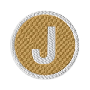 May include: Embroidered patch featuring the letter "J" in white, centered on a gold-colored circle. The circle is surrounded by a white border. The patch has a textured appearance, suggesting an embroidered design. The patch is circular in shape.