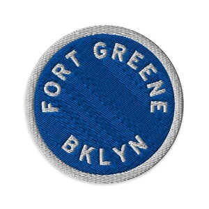 May include: A circular embroidered patch featuring a vibrant blue center with the white text FORT GREENE BKLYN stitched in a circular pattern. The patch is finished with a clean white merrowed border that frames the entire circular design.