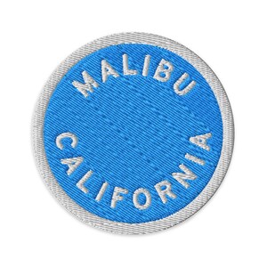 Könnte beinhalten: Ein kreisförmiger gestickter Aufnäher mit einer leuchtend blauen Mitte und dem weißen Text MALIBU CALIFORNIA, der entlang der oberen und unteren Kanten gebogen ist. Der Aufnäher hat einen dicken, weiß gestickten Rand.