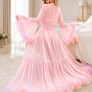 May include: A floor-length, sheer light pink robe featuring long, flared bell sleeves and a tiered skirt. The hem of the sleeves and the bottom of the robe are trimmed with a voluminous, multi-toned pink and lavender feather-like fringe detail.