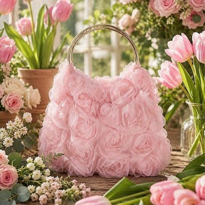 May include: A light pink handbag covered in layered fabric rosettes, featuring a circular gold-tone metal handle. The bag has a soft, textured appearance created by the overlapping petal-like fabric layers that form a dense floral pattern.