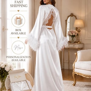 May include: A floor-length white satin robe featuring long sleeves with white feather trim at the cuffs and a back cutout detailed with white lace. The robe includes a matching waist tie and is shown alongside a gift box with the text For You.