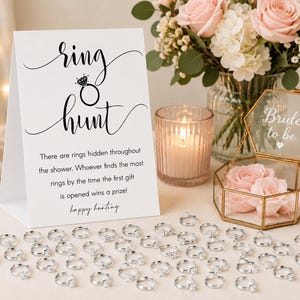 May include: A white tent-fold sign with black text reading ring hunt and instructions for a game, accompanied by a gold-rimmed glass box containing pink roses and the text bride to be, plus numerous small silver-toned rings scattered on the surface.