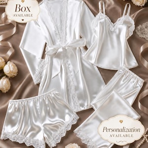 May include: A white satin pajama set consisting of a long-sleeve robe with lace trim, a camisole with lace detailing, shorts with lace hems, and long pants with lace hems. Text on the image includes Box AVAILABLE and Personalization AVAILABLE.