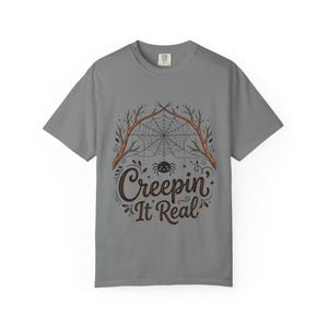 May include: A charcoal grey short-sleeve t-shirt featuring a graphic print of a spider hanging from a web between two bare tree branches. Below the spider, the text "Creepin' It Real" is written in a dark brown, stylised cursive script font.