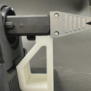 May include: A black mechanical sharpening tool attachment featuring a pointed, metallic, triangular tip with vertical ridges, supported by a white, angular, 3D-printed plastic stand that rests beneath the horizontal arm of the device.