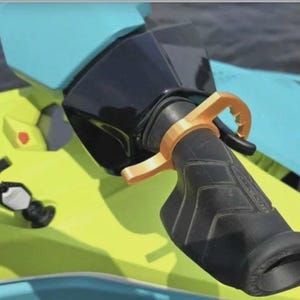 May include: A gold-coloured throttle assist lever attached to the black rubber handlebar grip of a personal watercraft. The watercraft features a bright lime green and light blue body with a black plastic housing near the handlebar assembly.