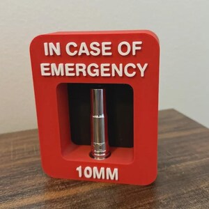 Può includere: Una cornice espositiva rettangolare rossa con il testo bianco IN CASE OF EMERGENCY in alto e 10MM in basso. Una singola bussola da 10 mm color argento è montata al centro della cornice su uno sfondo nero all'interno dell'apertura.