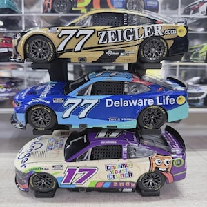 May include: Three stacked NASCAR diecast model cars. The top car is gold and black with the number 77 and Zeigler.com. The middle car is blue with the number 77 and Delaware Life. The bottom car is white and purple with the number 17 and Cinnamon Toast Crunch.