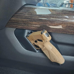 Puede incluir: Una pistola semiautomática de color tostado con empuñadura texturizada está montada en una funda de plástico negro a medida, integrada directamente en el panel interior gris oscuro de un vehículo, bajo un embellecedor de madera.
