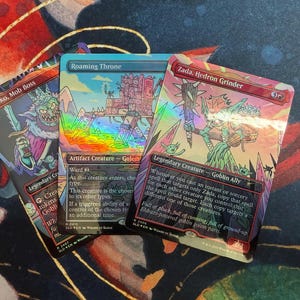 Może przedstawiać: Trzy karty kolekcjonerskie Magic: The Gathering. Karty przedstawiają kolorowe ilustracje fantastycznych stworzeń i krajobrazów. Nazwy kart to "Krenko, Mob Boss", "Roaming Throne" i "Zada, Hedron Grinder". Karty mają holograficzne wykończenie.