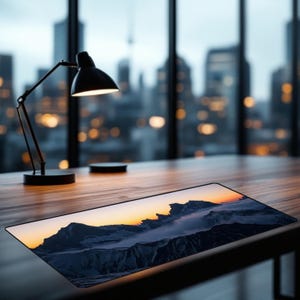 May include: A rectangular desk mat featuring a printed landscape of dark, snow-covered mountain peaks silhouetted against a vibrant orange and yellow sunset sky, with a layer of low-hanging clouds nestled between the jagged rocky summits.