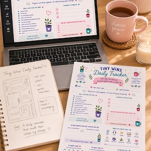 May include: A laptop displaying a Tiny Wins Daily Tracker digital template, a spiral-bound notebook with handwritten notes, a printed tracker sheet, a color palette card, a pink mug with the text choose peace, and sticky notes with motivational text.