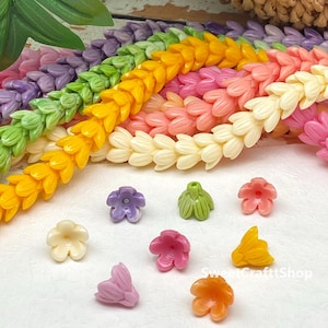 May include: A collection of small, multi-coloured flower-shaped beads in shades of purple, green, pink, yellow, cream, and orange. Several long, braided strands of these same flower beads are arranged in the background. The text SweetCraftShop is visible.