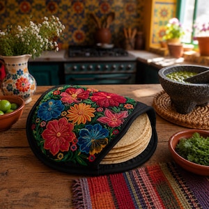 Op de afbeelding: Een zwarte tortilla warmer met een kleurrijk bloemenborduurontwerp, met een stapel tortilla's. De borduursels hebben rode, blauwe, oranje en roze bloemen. De warmer staat op een houten tafel met andere keukenartikelen.