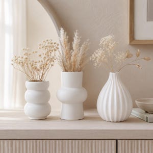 Peut inclure: Trois vases en céramique blanche de formes variées contenant des fleurs séchées. Le vase de gauche a une silhouette à trois bulles, celui du centre présente une forme cylindrique lisse avec un renflement, et celui de droite est côtelé.
