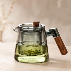 Può includere: Una teiera in vetro trasparente con una tonalità grigio scuro, dotata di un manico cilindrico in legno e un pomello in legno abbinato sul coperchio. Un infusore in vetro contiene foglie di tè verde immerse nel tè liquido.
