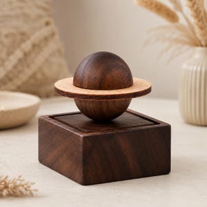 Peut inclure: Une sphère en bois brun foncé avec un anneau en bois beige clair autour de son centre, reposant sur une base carrée en bois brun foncé. La sphère est divisée en deux moitiés par l'anneau, créant une forme de planète sur un socle massif.
