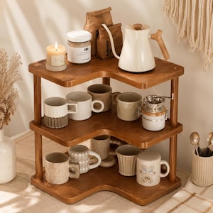 May include: A three-tiered wooden corner shelf unit featuring a warm brown finish. The top tier holds a white gooseneck kettle, a jar, and a lit candle. The lower tiers are filled with various ceramic mugs and a small metal coffee dripper.